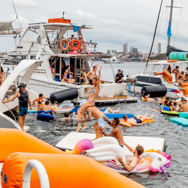 A lively group of people enjoy a summer day on the water, swimming and lounging on colorful inflatable floats, including a unicorn. Some people are on boats in the background from The Yacht Social Club Sydney Boat Hire, while others splash and play in the water near the dock.