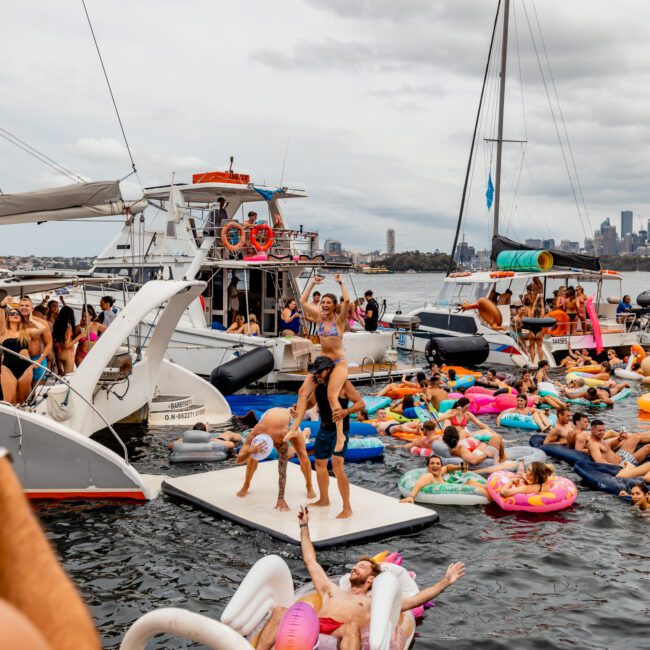 A lively scene of people enjoying a boat party on the water. Several boats from Sydney Harbour Boat Hire The Yacht Social Club are anchored, with guests swimming and relaxing on colorful inflatable floats. Some are dancing on a floating platform as the city skyline creates a picturesque backdrop.