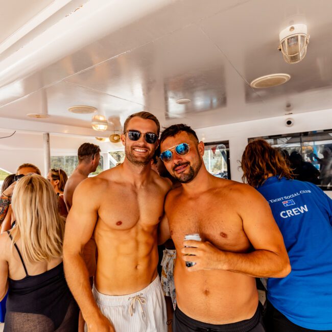 A group of people are on a boat enjoying themselves. Two shirtless men in the foreground, one with sunglasses, smile at the camera. A woman in a blue shirt with "CREW" printed on the back is also visible. Others in swimwear are socializing in the background, all part of The Yacht Social Club Sydney Boat Hire experience.