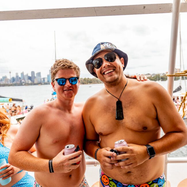 Two men wearing sunglasses and swimsuits are standing on a boat, holding drinks and smiling at the camera. The background shows a waterfront scene with boats and a city skyline under a cloudy sky, making it the perfect day for Sydney Harbour Boat Hire The Yacht Social Club.