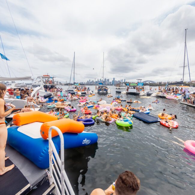 A lively scene of people enjoying a yacht social event on the water. Dozens of participants float on colorful inflatables near several anchored boats. The city skyline is visible in the background under a partly cloudy sky, showcasing the best of Luxury Yacht Rentals Sydney by The Yacht Social Club.