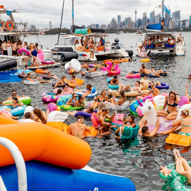 People are enjoying a sunny day on the water, using colorful inflatable floats near anchored boats. The crowd is lively, some sunbathing and socializing. The Sydney skyline is visible in the background. The scene is festive with various shapes and characters on the floats, embodying the spirit of Boat Parties Sydney The Yacht Social Club.