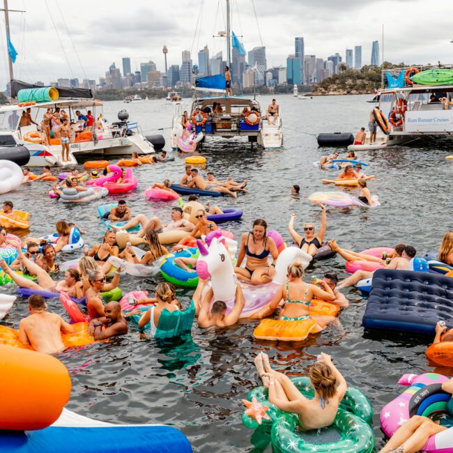 A lively group of people enjoying a floating party on inflatables in a waterway, surrounded by anchored boats. The background showcases a city skyline with tall buildings. Colorful inflatables, including a unicorn and pool floats, create a festive atmosphere reminiscent of The Yacht Social Club events.