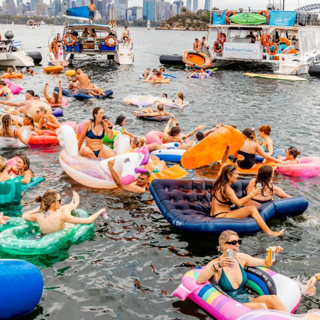 A vibrant scene of people enjoying a floating party on Sydney Harbour, surrounded by inflatables of various shapes and colors. Boats are docked nearby and a city skyline is visible in the background. The atmosphere is lively and festive, with people swimming and relaxing at The Yacht Social Club Event Boat Charters.