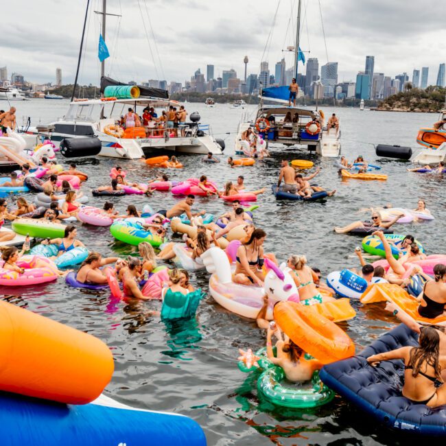 A lively scene of people enjoying a floating party in a harbor, with colorful inflatables, including a flamingo and a doughnut. Several boats from Boat Parties Sydney The Yacht Social Club are anchored nearby under the cloudy sky with a city skyline in the background. The atmosphere is festive and vibrant.