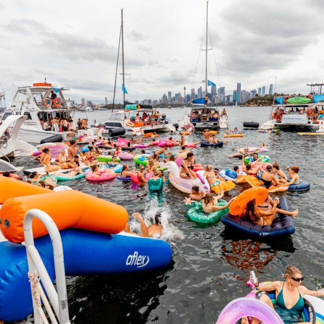 A lively scene of people enjoying The Yacht Social Club's boat party on the water. Many are floating on colorful inflatable rings and rafts. Several boats are anchored nearby, and a city skyline is visible in the background under a cloudy sky. The atmosphere is festive and crowded.