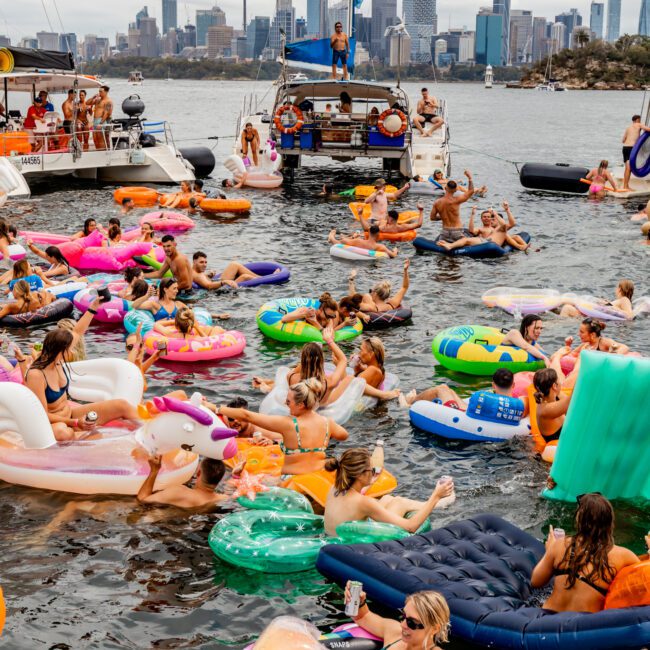 A lively scene on a sunny day with people enjoying The Yacht Social Club Event on the water. Many are lounging on colorful inflatables, including unicorns and doughnuts, near docked yachts. The background features a city skyline with tall buildings and a tower.