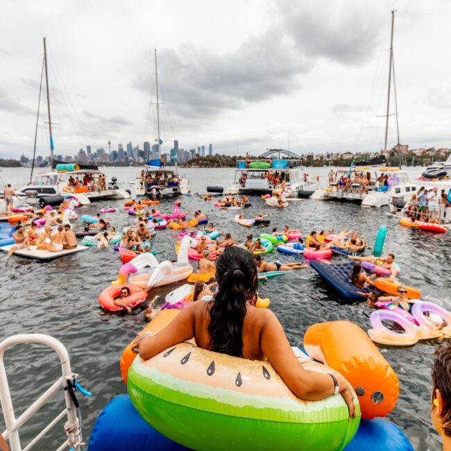 People enjoy a lively gathering on the water, floating on various inflatable rafts and loungers between yachts from The Yacht Social Club. The scene is vibrant and festive, with many participants socializing and relaxing. The city's skyline is visible in the background under a cloudy sky.