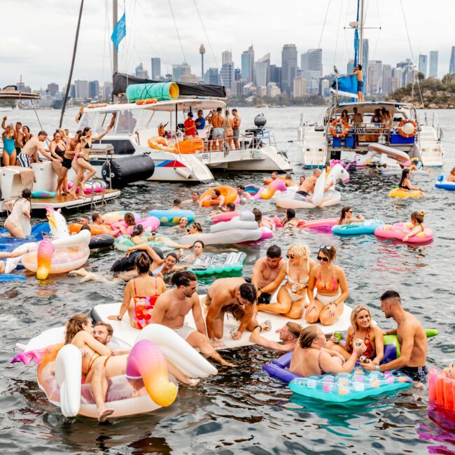 A lively party scene on the water features numerous people enjoying themselves on colorful floats and boats. The backdrop includes a city skyline with high-rise buildings. A mix of sailboats, motorboats, and luxury yacht rentals from The Yacht Social Club Sydney Boat Hire are anchored nearby, filled with more revelers.