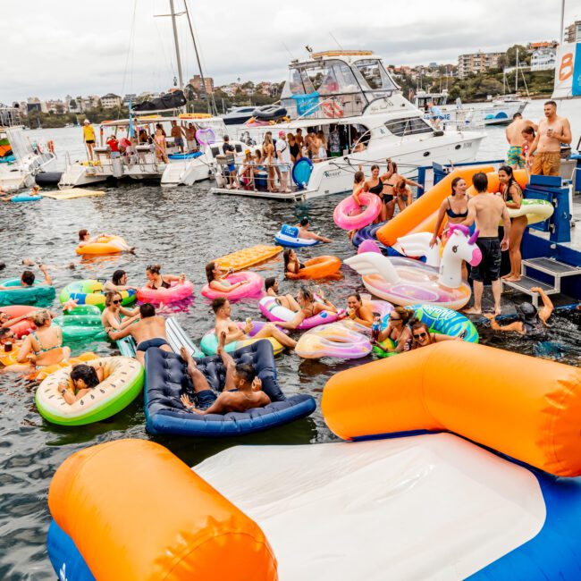 A lively scene of people on inflatable rafts and pool floats in the water near boats, socializing, swimming, or lounging. The background features parked yachts and a cityscape. The atmosphere suggests a festive summer gathering on the water, typical of events hosted by The Yacht Social Club Sydney Boat Hire.