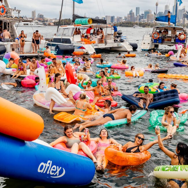 A joyful crowd enjoys a party on a lake, floating on colorful inflatable rafts and pool floats, including unicorns, flamingos, and pizza slices. Behind them are several docked boats from The Yacht Social Club Sydney Boat Hire, with a city skyline visible in the background.