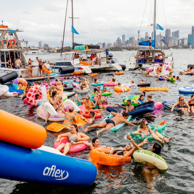 A lively scene on a river with numerous people on colorful inflatables, including unicorns and flamingos, surrounded by boats. The city skyline is visible in the background. The atmosphere is cheerful and festive as people relax and enjoy the water, reminiscent of The Yacht Social Club Sydney Boat Hire.