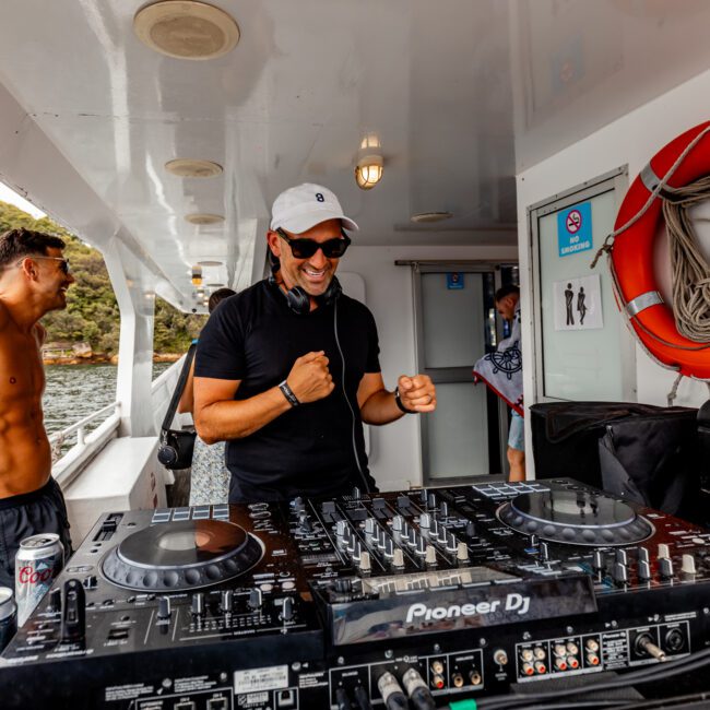 A DJ wearing sunglasses and a white cap is smiling and playing music on a boat, with DJ equipment in front of him. A shirtless man stands nearby, leaning on the railing. The scene takes place on a boat with water and a forested backdrop, showcasing The Yacht Social Club Event Boat Charters.