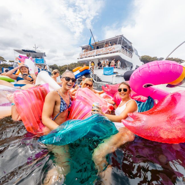 A group of people enjoys a sunny day on the water with large inflatable floats shaped like flamingos and mermaids. They smile and laugh near a boat named "Serenity Explorer," set against blue skies and trees, showcasing Boat Parties Sydney The Yacht Social Club.