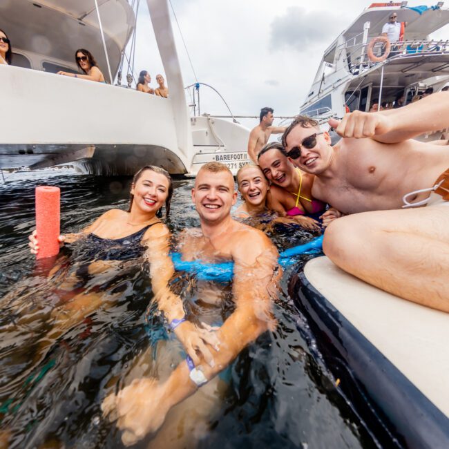 A group of friends enjoys a swim and relaxation in the water, next to a boat. They are all smiling and appear to be having a great time. Boats and other people can be seen in the background, indicating a lively atmosphere typical of Sydney Harbour Boat Hire The Yacht Social Club.