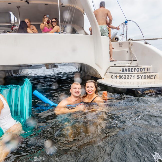 A couple is happily swimming in the ocean near the back of a luxury yacht named "Barefoot II" with registration "ON-862271 SYDNEY". Other people are on the boat, some taking photos. The atmosphere appears joyous and fun, as if part of a Boat Parties Sydney event. The water is slightly splashing up in the foreground.