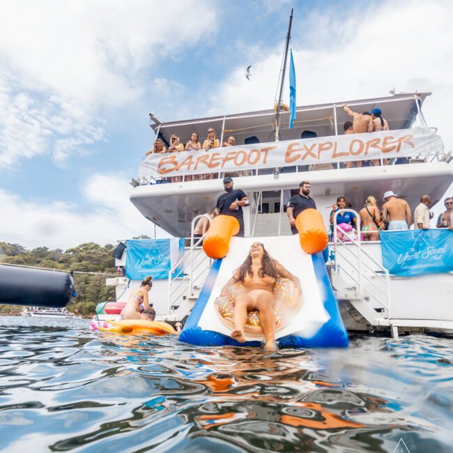 A party boat named "Barefoot Explorer" is anchored on water, filled with people. One person is sliding down an inflatable slide into the water. Others are lounging on the boat and inflatable tubes. The setting appears sunny and lively, a perfect day for Luxury Yacht Rentals Sydney with a scenic backdrop in Sydney Harbour Boat Hire.