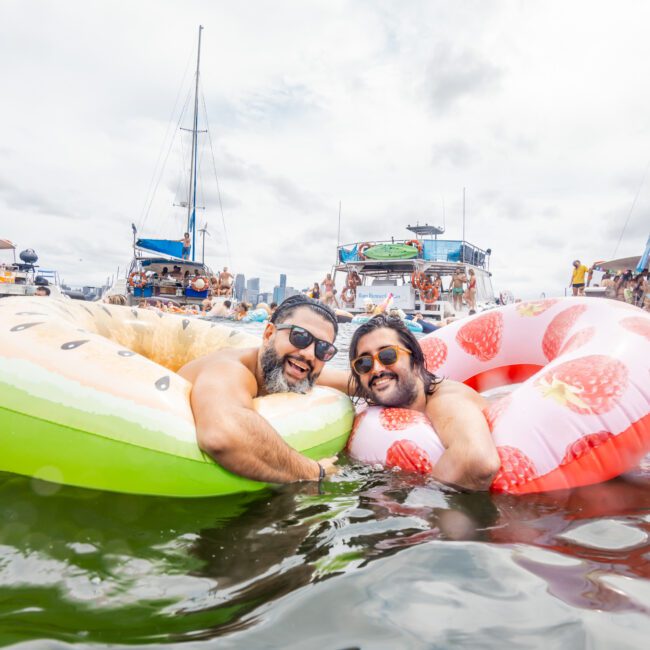 Two smiling individuals in inflatable fruit-themed pool floats (one is a kiwi slice, the other a strawberry) enjoy a day on the water. Boats and people in the background suggest a bustling, social environment on a partly cloudy day. Text on the image reads "The Yacht Social Club Event Boat Charters.