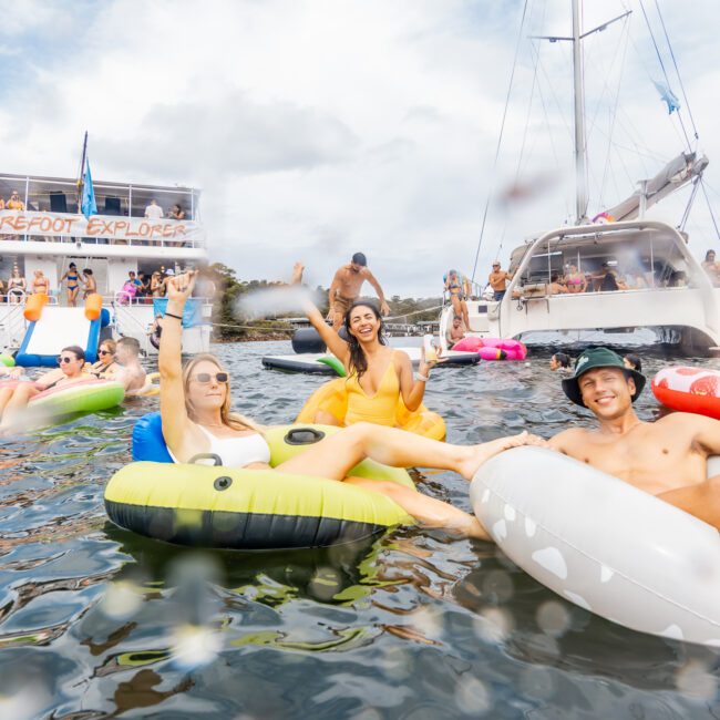 A group of people are enjoying a sunny day on the water, floating on various inflatable tubes and boats. Some are drinking and raising their hands while others relax. A large yacht and a houseboat labeled "Barefoot Explorer" are docked in the background, part of The Yacht Social Club's Luxury Yacht Rentals Sydney.