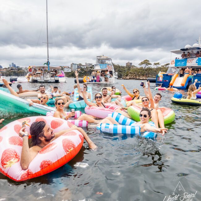 A group of people relax on colorful inflatable pool floats in the water near several boats. They are smiling, holding drinks, and enjoying a festive atmosphere under a cloudy sky. Some of the boats, from Luxury Yacht Rentals Sydney, have slides and decorations, adding to the lively scene.