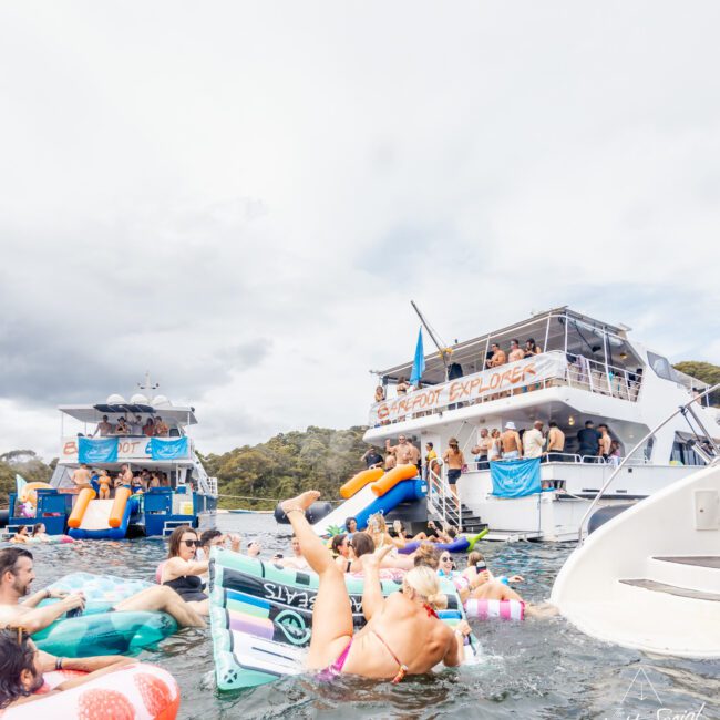 A lively boat party with guests swimming and floating on inflatables in the water. Two boats from Sydney Harbour Boat Hire's Yacht Social Club are docked nearby, with partygoers both on board and in the water. The atmosphere is festive, featuring colorful decorations and banners against a cloudy sky.