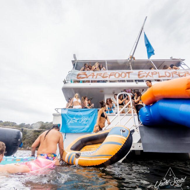 A group of people are having a lively party on a large boat named "Barefoot Explorer." Some are swimming near the boat, while others are on the deck. Various inflatable tubes are visible. The scene is festive with a "The Yacht Social Club Sydney Boat Hire" banner in the background.