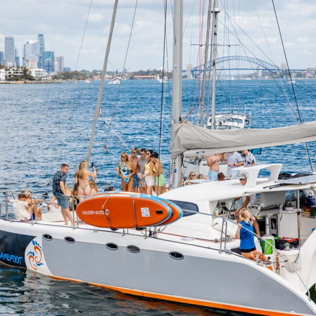 A group of people enjoy a sunny day on a sleek, modern yacht anchored in a calm bay with city skyscrapers visible in the background. The yacht features a vibrant orange kayak on deck. Some people stand, others sit or lounge, all dressed for warm weather.