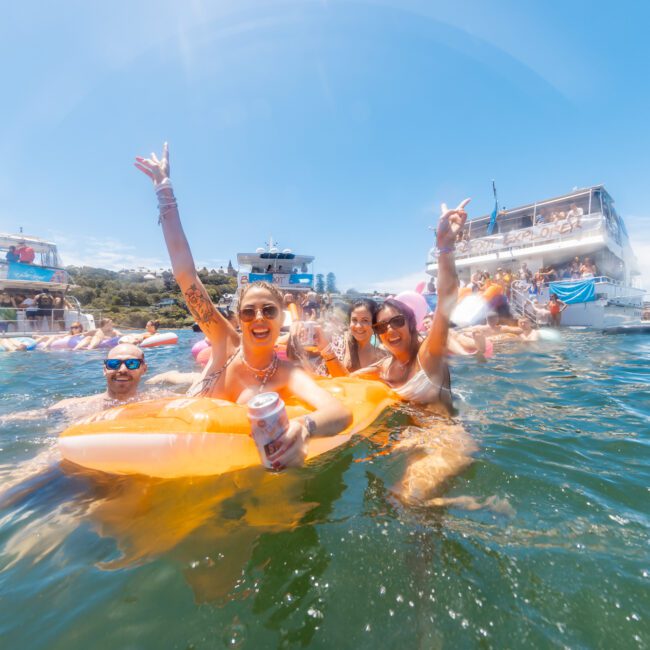 A lively group of people enjoys a sunny day at the lake, surrounded by large party boats. Some are floating on inflatables, holding drinks, and making celebratory hand gestures. The atmosphere is festive, with clear skies and lush greenery in the background.