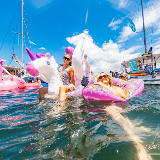 Two women relax on inflatable unicorns during a sunny day. One sits on a white unicorn float, and the other lounges on a pink one. Boats and other people are seen in the background, under a blue sky with scattered clouds, creating an idyllic summer scene.