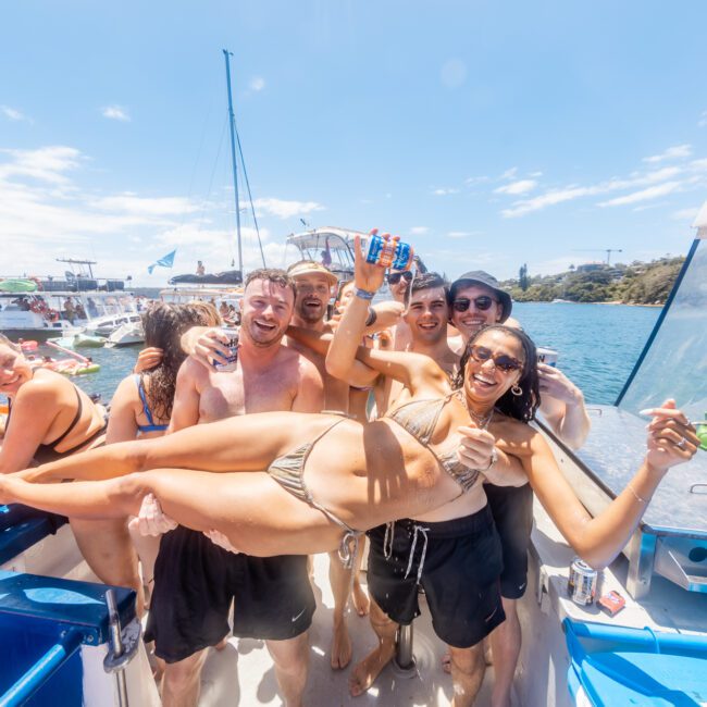 A group of friends on a boat having fun under a clear blue sky. One person, wearing a bikini, is being playfully hoisted by others. They are smiling and posing for the camera, enjoying a sunny day on the water with yachts dotting the coastline in the background.