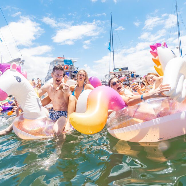 A group of people enjoying a sunny day in the water, floating on large inflatable unicorns and other colorful floats. Sailboats gracefully glide in the background as everyone smiles and has fun. The image captures a lively, festive atmosphere with an unmistakable summer vibe.