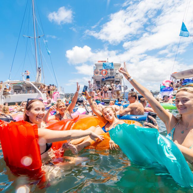 A group of smiling people in swimsuits, floating on colorful inflatable loungers in the water, with several boats filled with more people in the background. The clear blue sky indicates a sunny day. Everyone appears to be enjoying a lively beach party under perfect conditions.