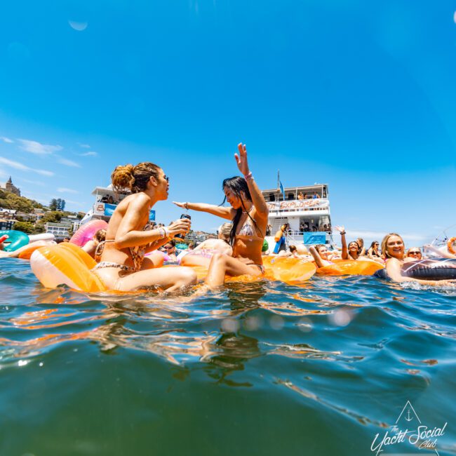 Two women in bikinis enjoy a lively party on an inflatable in the water, with a multi-deck yacht in the background. Surrounded by other partygoers under a clear blue sky, the scene is festive and energetic. The "Yacht Social Club" logo is visible on multiple inflatables.