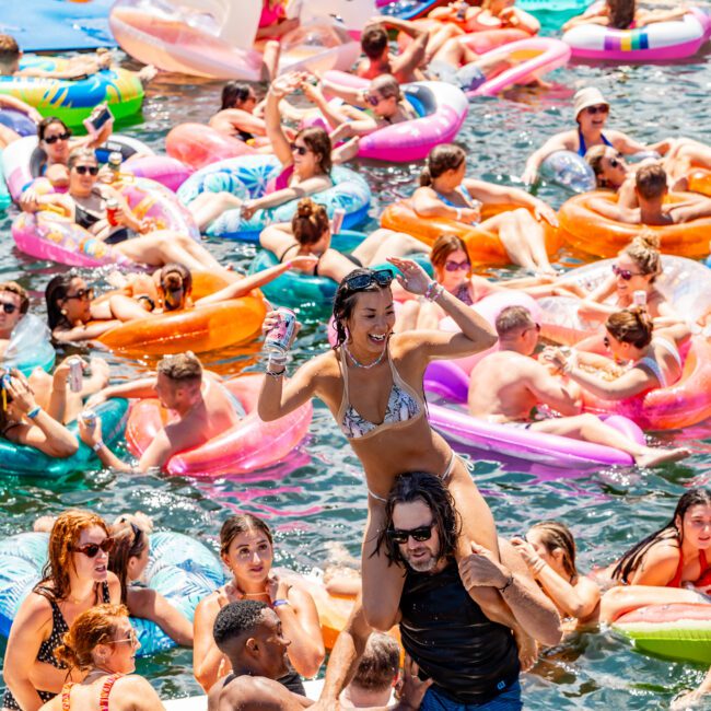 A crowded pool party with numerous people enjoying themselves on colorful inflatable floats, including unicorns and flamingos. In the foreground, a man carries a woman on his shoulders while she holds a sparkling drink and raises her arm in excitement.