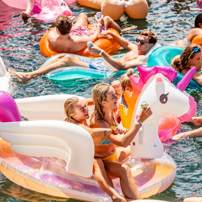 People enjoying a sunny day on the water, riding various inflatable floats including a unicorn float. Some are holding drinks and taking selfies on their phones. The water is filled with colorful inflatables while everyone relaxes and has fun.