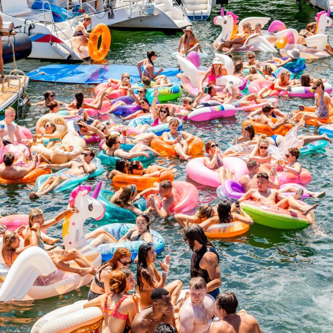 A lively scene features a large group of people enjoying a sunny day on the water. They are floating on colorful inflatable rafts, including unicorns and donuts. Paddleboarders and boats in the background add to the festive, crowded atmosphere.