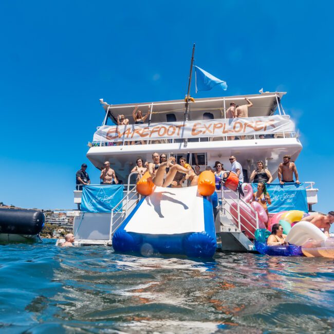 A lively scene of people enjoying a sunny day on a boat labeled "Barefoot Explorer," equipped with a water slide. Some are sliding into the water, while others relax on colorful floating inflatables. Scenic cliffs and charming houses appear in the background, creating a picturesque setting.