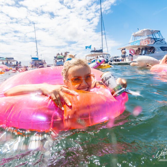 A person relaxes in a pink inflatable tube while holding a canned drink, surrounded by people and boats on a sunny day. The sky is clear and the water is calm. In the background, kayaks and swimmers can be seen enjoying the scene.