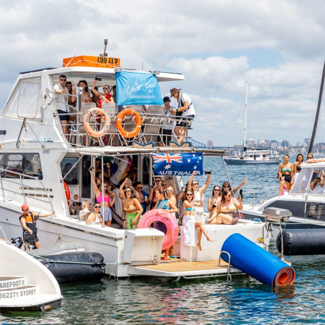 A group of people is gathered on a multi-level boat, enjoying a sunny day on the water. The boat is decorated with inflatable pool toys and flags, including an Australian flag. The harbor is visible in the background. Some participants are in swimwear, lounging on a floating mat in the water.