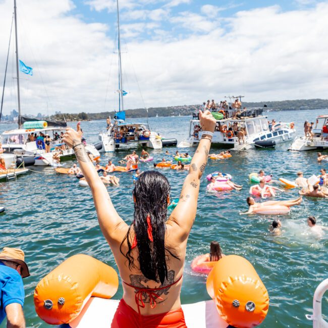 A woman with long hair and a red bikini stands on a boat, raising her hands in the air while looking at multiple yachts and a crowd of people swimming and lounging on inflatables in the water during a sun-drenched day.