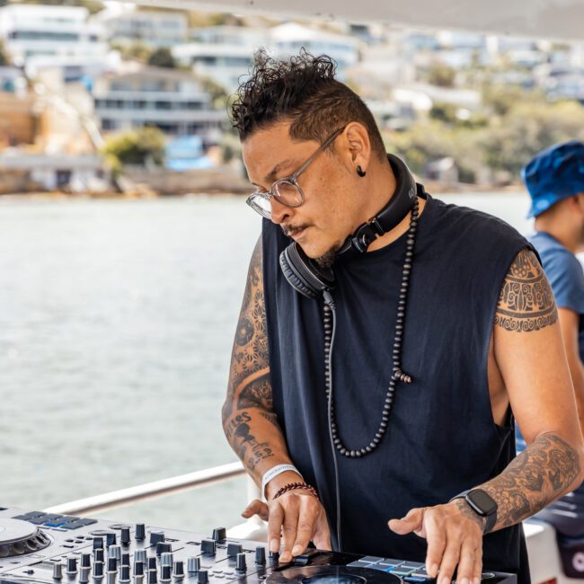 A DJ with tattoos and glasses is leaning over his equipment, wearing headphones around his neck. He is on a boat with waterfront buildings and trees in the background. Another person wearing a blue hat is partially visible beside him, adding to the scene's lively atmosphere.