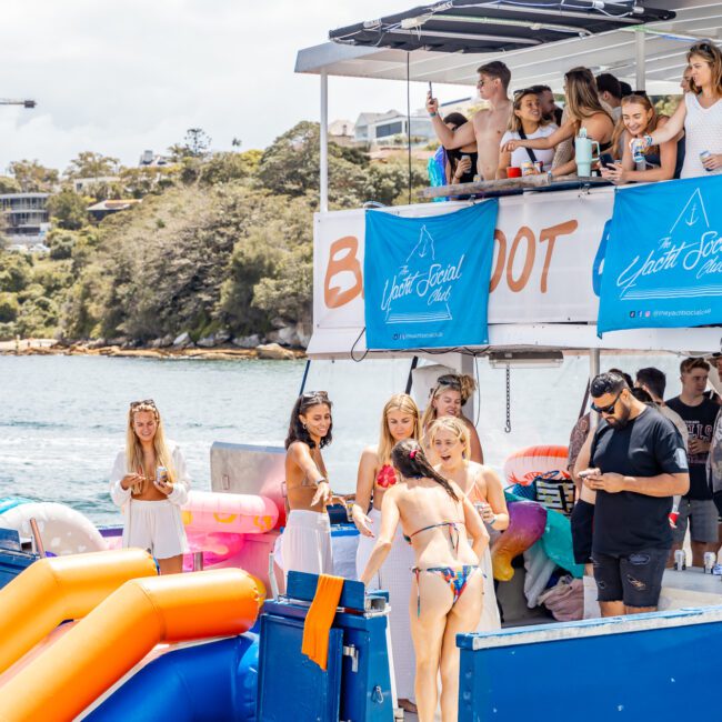 A group of people are enjoying a sunny day on a yacht. Some relax on the upper deck while others gather near the waterslide on the lower deck. The yacht features festive banners for an event and is docked, with a green hilly shore visible in the background, creating an idyllic setting.