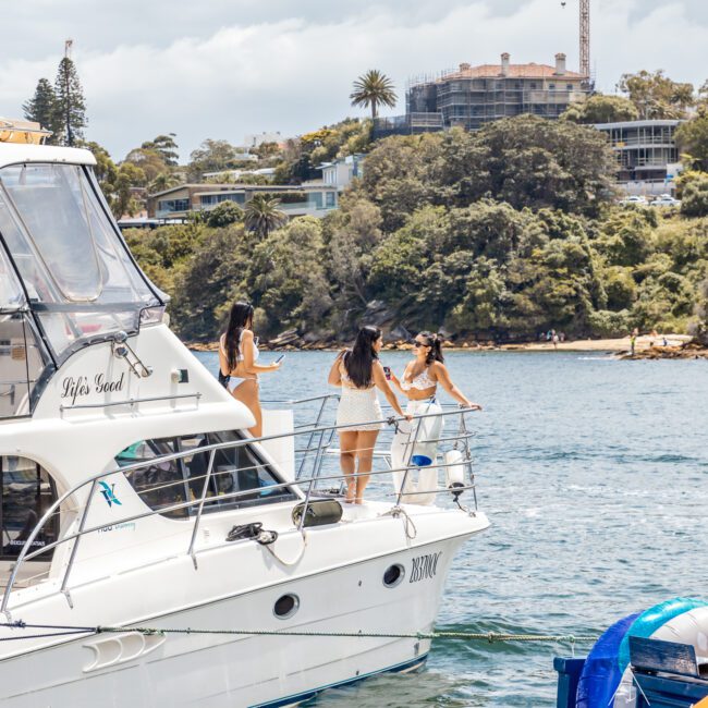 Three women are on the deck of a yacht named "BUBBLY," enjoying a sunny day on the water. They appear to be in conversation. In the background, there's a shoreline with trees, buildings, and a crane at a construction site. The yacht proudly displays the "Yacht Social" logo on its side.