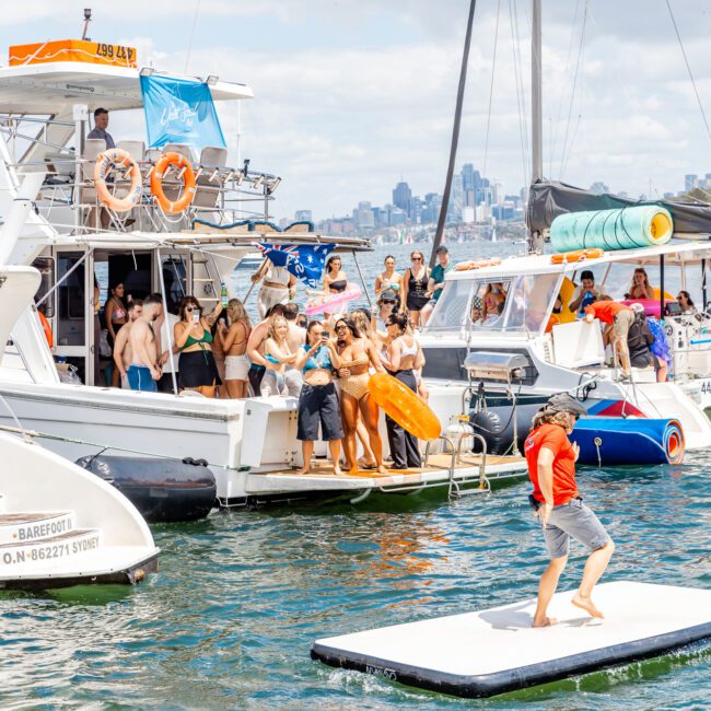 A lively scene on the water with a group of people partying on a docked boat. Some individuals are dancing, while others are socializing aboard the boat. Another person is in the foreground on an inflatable platform, surrounded by other boats and with a city skyline creating a picturesque backdrop.