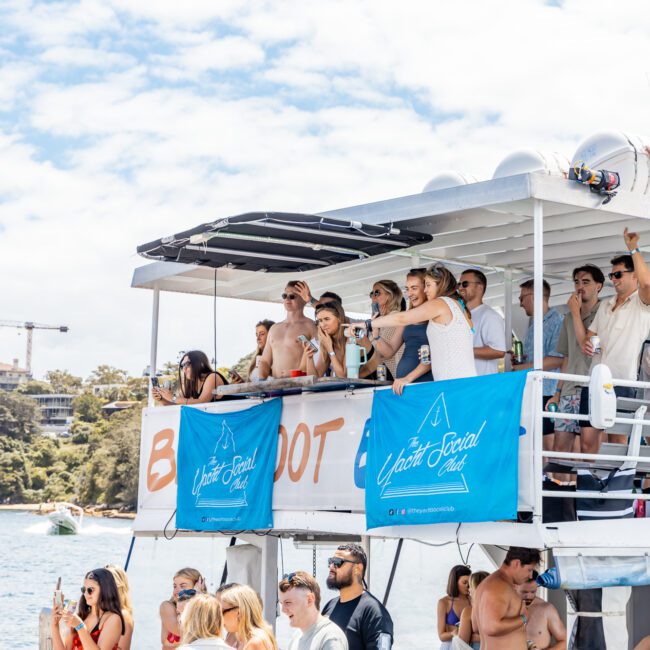 A group of people enjoys a sunny day on a two-story boat, the "Barefoot Boat." Many are standing on the upper deck, shaded by a canopy, and a few are on the lower deck. Blue banners with "The Yacht Social Club Event Boat Charters" are displayed. The background shows water and greenery.