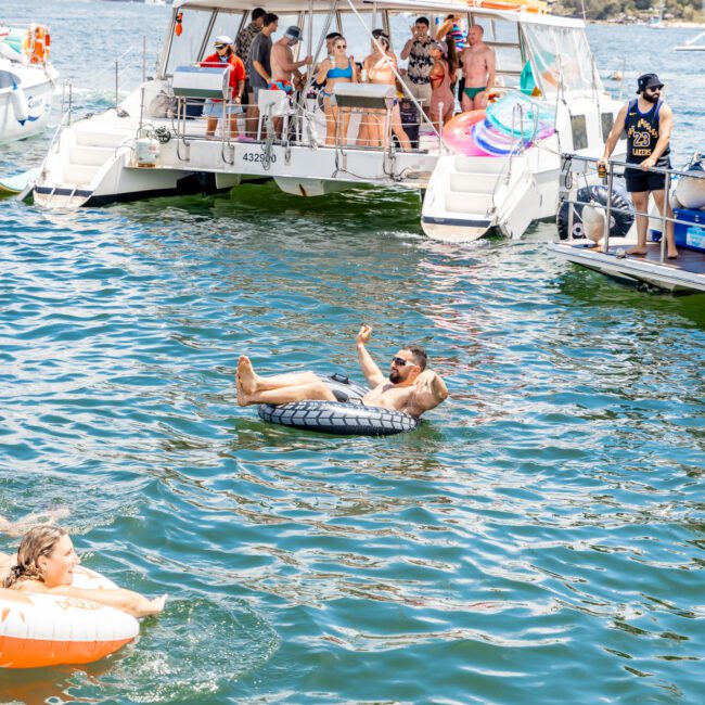 A group of people enjoys a sunny day on a luxurious catamaran. Some are on the boat, others float in the water on inflatable rings, and a woman swims nearby. The background reveals a tree-covered shoreline under a partly cloudy sky.