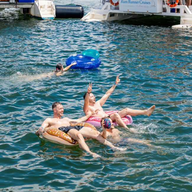 Three people float on inflatable swim rings in the water, enjoying a sunny day. One raises a peace sign while another nearby swimmer wears goggles and splashes water. Boats with more people onboard are anchored in the background, while music plays, adding to the lively and festive atmosphere.