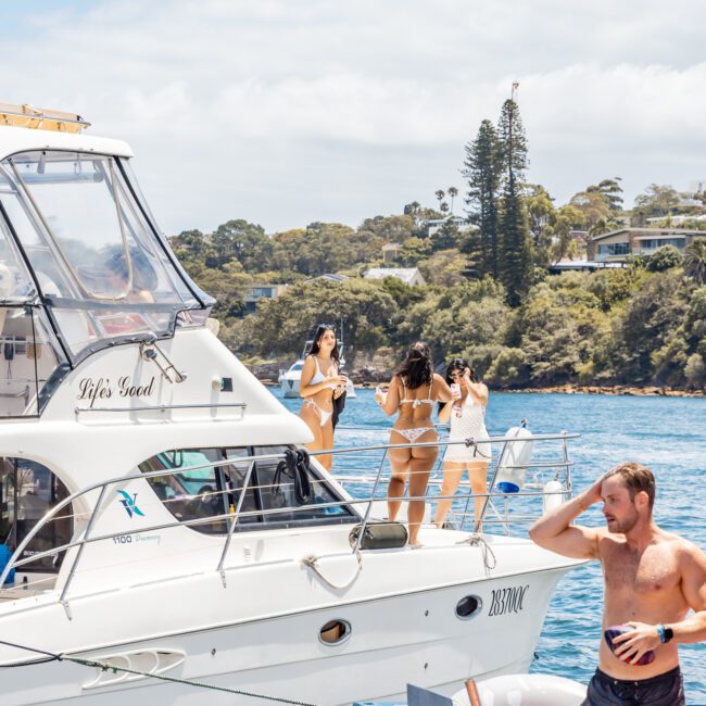 A group of people enjoying a sunny day on a yacht named "Life's Good" docked next to another boat on the water. Two women in swimsuits stand on the deck, while a shirtless man looks towards the water from an orange inflatable raft nearby. A jet ski zooms by, adding excitement to the scene with trees in the background.