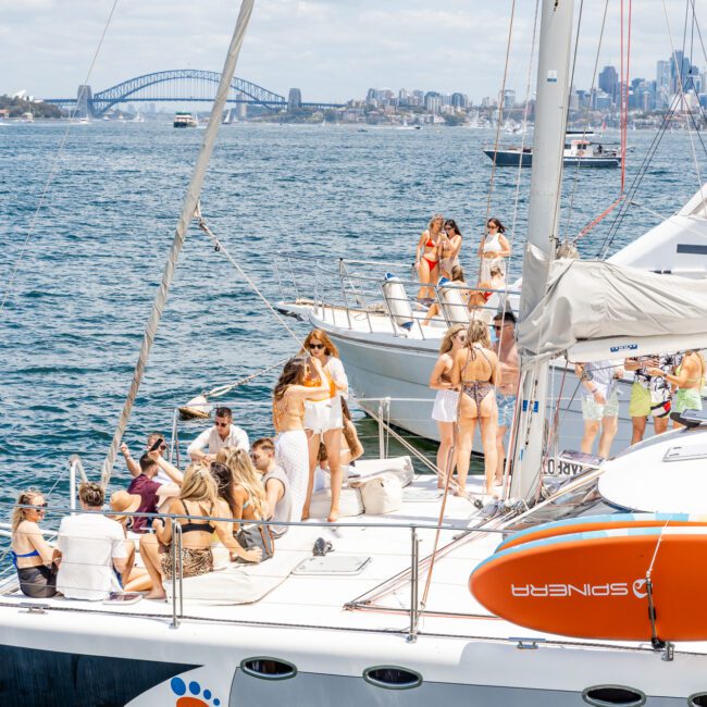 A group of people socialize on a yacht anchored in a bay with a picturesque view of a city skyline and a prominent arch bridge in the background. The weather is sunny, and everyone appears to be enjoying the outdoor gathering under clear skies.