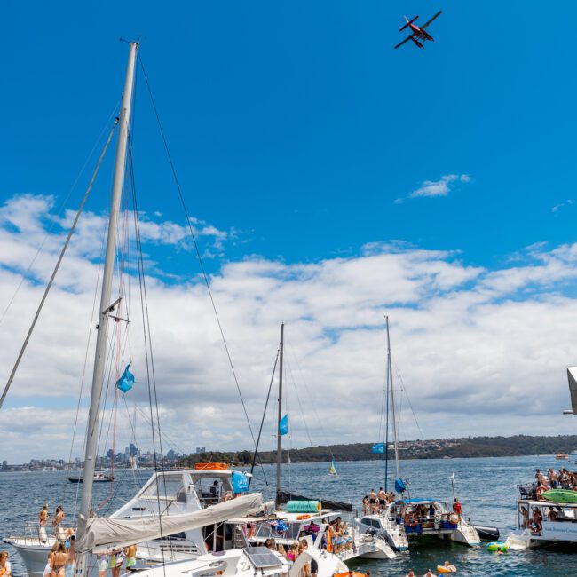 A lively scene of several sailboats and yachts on a sunny day with numerous people socializing and relaxing. The sky is clear with a few clouds, and a small plane is flying overhead. People are also seen floating on colorful inflatables in the water, adding to the festive atmosphere.